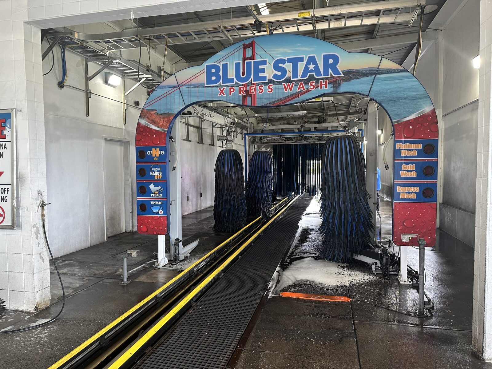 Looking out from inside the Blue Star Xpress Wash express tunnel toward the daylight exit, with yellow guide rails along the track